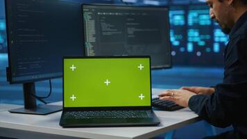 Isolated screen notebook in front of employee working in server farm used for processing data. Close up shot of mockup laptop on desk next to IT developer typing at PC, updating servers video