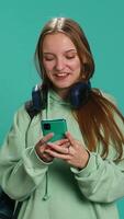 vertikal porträtt av Lycklig tonåring läsning meddelanden på telefon, förtjust efter tar emot Bra Nyheter. glad kvinna upphetsad förbi SMS på smartphone, firar, studio bakgrund, kamera en video