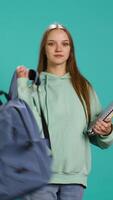 Vertical Portrait of smiling woman with backpack holding school notebook and notes, isolated over studio background. Cheerful girl with school supplies in arms, preparing to go to university, camera B video