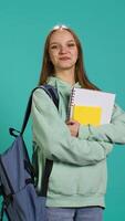 Vertical Portrait of joyous teenager with rucksack holding school notebook and notes, isolated over studio background. Jolly pupil with school supplies in arms, preparing to go to college, camera B video