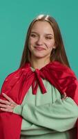 Vertical Radiant woman grinning while portraying confident superhero wearing red cape, isolated over studio backdrop. Portrait of jolly heroic person posing as powerful hero character in costume, camera A video