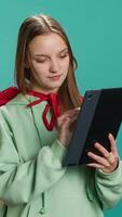 Vertical Young girl wearing superhero costume for Halloween browsing social media feed on tablet, studio background. Woman dressed as comic book hero having fun reading messages on device, camera A video