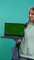 Vertical Happy girl presenting laptop with green screen display, isolated over studio backdrop. Radiant teenager creating promoting advertisement with mockup notebook device, camera B video