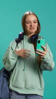 Vertical Cheerful woman saluting friends during teleconference meeting using smartphone, studio background. Teenager having fun catching up with school colleagues during videocall, camera B video