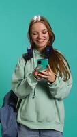 Vertical Portrait of happy teenager reading messages on phone, delighted after receiving good news. Cheerful woman excited by SMS on smartphone, celebrating, studio background, camera B video