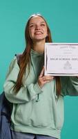 verticaal portret van glimlachen leerling Holding hoog school- diploma, vieren voorbijgaan klassen met hoog merken, Bijwonen diploma uitreiking ceremonie. gelukkig jong meisje verheugd over ontvangen certificering, camera b video