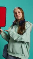 Vertical Smiling woman holding red speech bubble sign of empty copy space for message. Joyous teenager presenting thought bubble cardboard used as promotion concept, isolated over studio background, camera B video