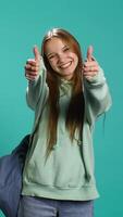 Vertical Happy girl doing thumbs up hand gesturing, showing approval, having positive mood. Smiling teenager presenting confirmation, doing agreement finger sign, studio background, camera B video