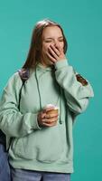 Vertical Young girl with backpack yawning, feeling tired, drinking coffee to wake up. Sleepy woman using caffeinated beverage to gain energy, lacking sleep, studio background, camera B video