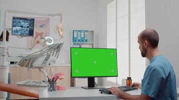 Man working with keyboard and computer with green screen on desk in dentistry office. Oral care assistant using mockup template and isolated background in dental cabinet for teethcare video