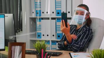 Entrepreneur wearing face protection against coronavirus using phone for videomeeting talking with remotely coworker sitting in new normal office corporate building. Woman respecting social distance video