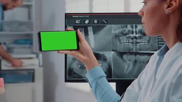 Close up of dentist analyzing smartphone with green screen and teeth scan on computer in dental cabinet. Woman holding device with chroma key for isolated mockup template for dentistry video