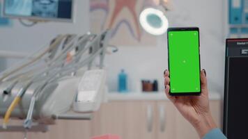 Close up of dentist holding smartphone with green screen for teethcare at dental cabinet. Dentistry specialist looking at chroma key and isolated mockup template on mobile phone. video