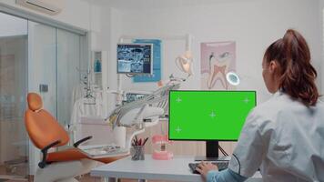 Woman working as dentist with green screen on monitor at dental cabinet while assistant preparing tools for teethcare. Specialist using computer with chroma key and mockup template video