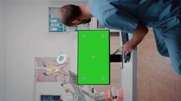 Vertical . Assistant using horizontal green screen on computer for dental care. Man working with mockup template and isolated background on monitor for teethcare at dentist office. video