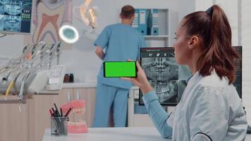 mujer trabajando con horizontal verde pantalla en móvil teléfono para cuidado de los dientes en dentista oficina. ortodoncista mirando a croma llave y Bosquejo modelo en teléfono inteligente para dental cuidado. video