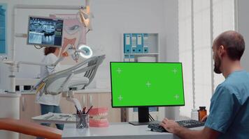 Assistant using horizontal green screen on computer while dentist preparing equipment for oral care examination in dental cabinet. Man looking at chroma key and mockup template on monitor video