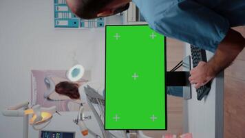 Vertical . Assistant looking at horizontal green screen on computer at dentist office. Man working as teethcare specialist with mockup background and isolated template on monitor video