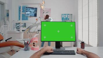 POV of man using keyboard and computer with green screen at desk in oral care cabinet. Assistant looking at monitor with isolated background and mockup template for teethcare and dental checkup video