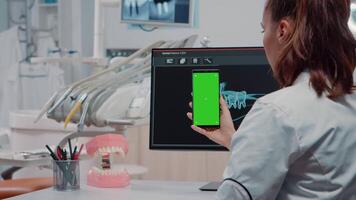 Dentist vertically holding smartphone with green screen while looking at teeth scan on computer in office. Teethcare specialist using mobile phone with chroma key for isolated background video