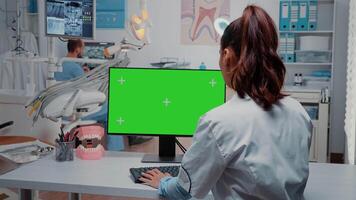 Dentist working with horizontal green screen on monitor at desk while talking to man assistant for dental care. Woman using computer with chroma key and isolated template for dentistry video