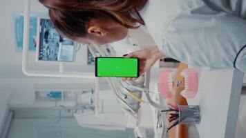 vertikal . kvinna vertikalt innehav mobil telefon med grön skärm i dental skåp. tandläkare ser på isolerat attrapp bakgrund med krom nyckel på visa för tandvård. video