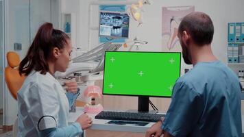 Oral care team analyzing teeth layout while using computer with horizontal green screen on desk. Dentist and dental assistant working with tools and equipment for dentistry and teethcare video