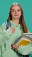 vertical retrato de sonriente mujer con mochila participación colegio cuaderno y notas, aislado terminado estudio antecedentes. alegre niña con colegio suministros en brazos, preparando a Vamos a universidad, cámara un video