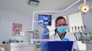 dentisterie médecin interroger patient et prise Remarques sur presse-papiers avant chirurgie. médecin et infirmière travail dans moderne orthodontique bureau, l'écriture et examiner la personne portant protection masque video