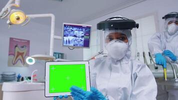 Patient pov of dentist in ppe suit explaining dental radiography and diagnosis for teeth infection using tablet with green screen. Stomatology specialist pointing at mockup, copy space, chroma display video