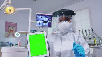 Doctor in ppe suit showing tablet with green screen, explaining dental radiography and diagnosis for teeth infection. Stomatology specialist in coverall pointing at mockup, copy space, chroma display video