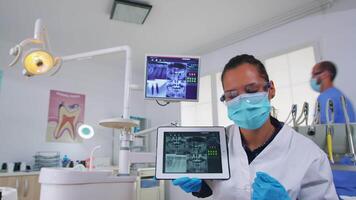 paciente pov dentro dental escritório discutindo tratamento do dentes cavidade, dentista apontando em digital raio X usando tábua. equipe do médicos trabalhando dentro moderno estomatológico clínica, explicando radiografia dente video