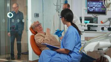 Zahnarzt Assistent verhören Senior Frau und nehmen Anmerkungen auf Zwischenablage warten zum Arzt. Senior geduldig im Schmerzen erklären Dental Problem zu Krankenschwester Anzeige beim Zahnschmerzen Probleme video