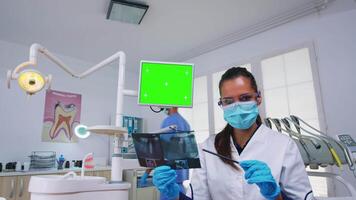 Dentist doctor showing unit monitor with mockup screen, holding teeth radiography and examining teeth infection. Stomatology specialist with face mask pointing at greenscreen copy space chroma display video