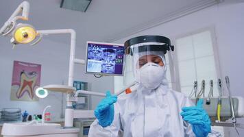 dentista punto de ver vistiendo proteccion engranaje en contra codicioso brote explicando tratamiento en dental oficina. estomatólogo vistiendo la seguridad engranaje en contra coronavirus durante cuidado del calor cheque de paciente. video