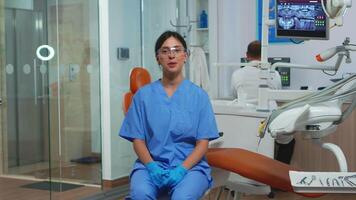 Dental assistant looking at camera speaking with patients about dental hygiene. Stomatologist talking on webcam sitting on chair in stomatological clinic with doctor in background working on computer video