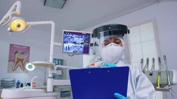 Pov of patient getting dental treatment in dentist office with new normal, doctor taking notes on clipboard before examining. Stomatolog wearing safety gear against coronavirus during heatlhcare check video