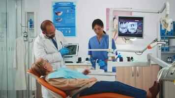 Dentist showing on tablet teeth x-ray reviewing it with patient. Doctor and nurse working together in modern stomatological clinic, explaining to old woman radiography of tooth using notebook display video