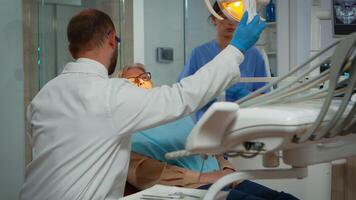 Orthodontist lighting the lamp until examination and patient opening mouth. Stomatologist speaking to woman with toothache sitting on stomatological chair while nurse preparing tools for surgery. video