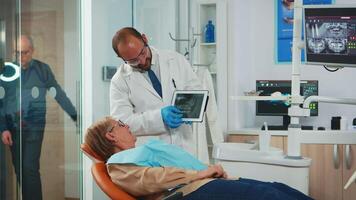 Stomatologist reviewing x ray of tooth with senior patient explaining treatment. Dentist showing to old woman dental radiography using tablet, doctor and nurse working together in modern clinic. video