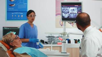 Man dentistry doctor pointing on digital screen showing dental implants. Dentist and nurse working together in modern stomatological clinic, explaining radiography of teeth on monitor video
