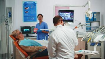 Stomatologist explaining proper dental hygiene using presentation teeth skeleton. Dentist telling to senior patient procedure holding sample of human jaw with a toothbrush in stomatology office. video