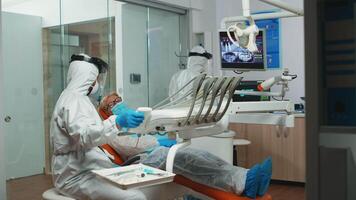 dentista técnico dentro macacão preparando para dental cirurgia iluminação a luminária dentro estomatológico escritório durante global pandemia. médico equipe vestindo proteção terno, face escudo, mascarar e luvas. video