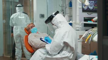 tandläkare sjuksköterska i skyddande kostym tar anteckningar på Urklipp tala med senior patient under coronavirus pandemisk. begrepp av ny vanligt ortodontist besök i coronavirus utbrott bär skyddande kostym och ansikte skydda video