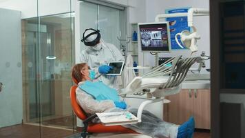 Dentist in protective equipment showing on tablet dental x-ray reviewing it with senior patient. Medical team wearing face shield coverall, mask, gloves, explaining radiography using notebook display video