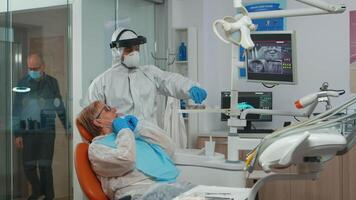 Doctor with protective mask doing hygienic cleaning of teeth using sterile dental tools during coronavirus pandemic. Medical team wearing coverall, face shield, mask, gloves in stomatological clinic video