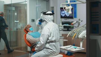 Stomatologist in protective suit asking for patient dental x-ray examining teeth problems during coronavirus pandemic in modern clinic. Medical team wearing coverall, face shield, mask and gloves. video