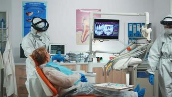 Dentist wearing protection equipmentin dental office examining x-ray image on tablet during global pandemic. Medical team with face shield, coverall, mask reviewing radiography using digital device video