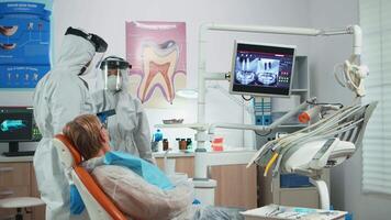 Dentist with face shield reviewing panoramic mouth x-ray image to patient during global pandemic. Assistant and doctor talking with senior woman wearing suit, coverall, protection suit, mask, gloves video