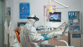 Dentist with face shield working in dental unit during covid-19 pandemic using sterilized tools. Orthodontist and nurse working wearing coverall, face shield, mask, gloves in stomatological office video